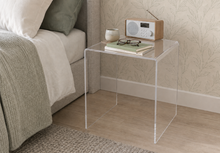 A clear acrylic side table placed beside a bed in a softly styled bedroom. The transparent table holds stacked books, a small ceramic cup, eyeglasses, and a light wood-and-white radio.