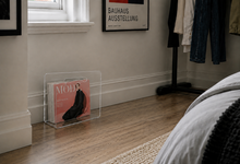 A clear acrylic magazine rack holding a fashion magazine on a wood floor in a styled bedroom corner with a bed, wall art, and clothing rack visible.