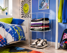 Two clear acrylic stackable storage shelves holding folded T-shirts and sneakers in a bright bedroom setting.