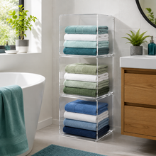 A stack of three clear acrylic storage shelves filled with folded towels in shades of blue, green, and white, set in a bright, modern bathroom next to a bathtub and wooden vanity.